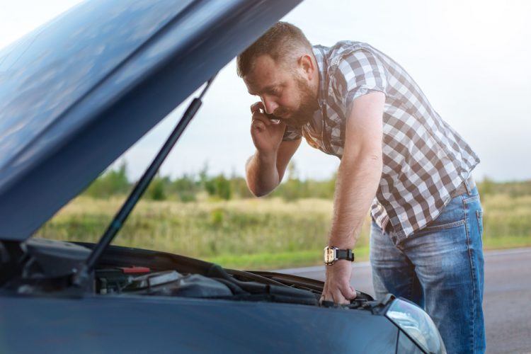 man with a phone in front of the open hood of a broken car