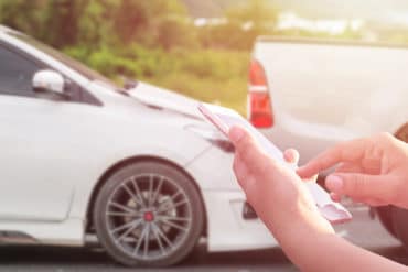 Close up hand of woman using smartphone and blur of her broken car parking on the road. Contacting car technician, Insurance or need help concept