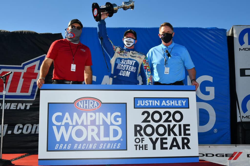 Justin Ashley celebrates winning the 2020 NHRA Rookie of the Year with one person on each side, standing in front of a podium